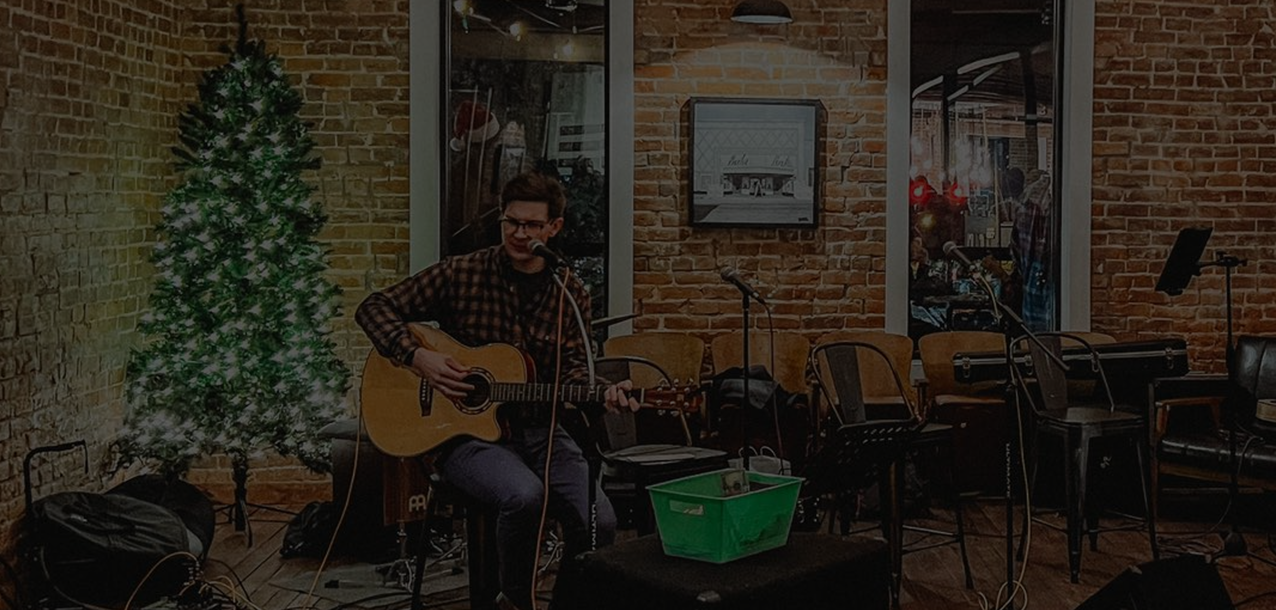 A man with glasses and a flannel shirt sitting on a barstool with a guitar, singing into a microphone at Rapp's Barren Brewing company in Mountain Home Arkansas offers, darkened for the website header page
