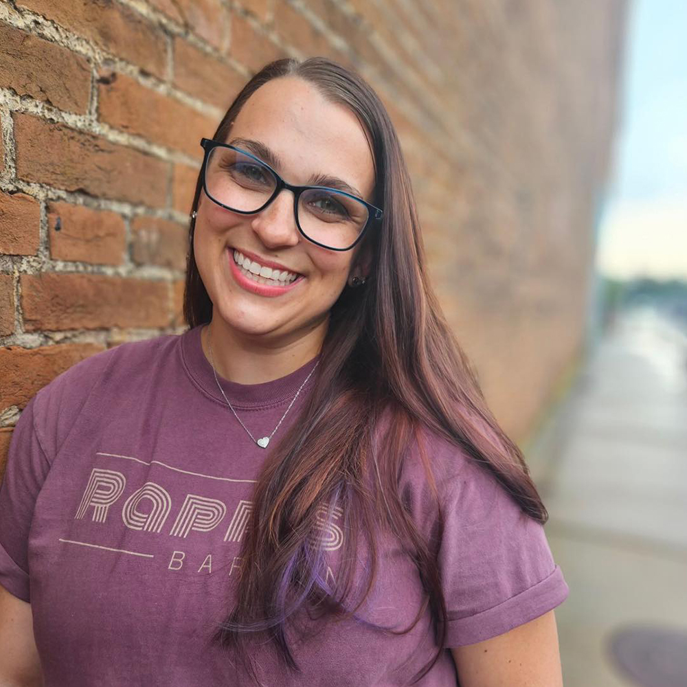 Beautiful smiling woman with glasses standing next to a brick wall wearing a Rapp's Barren t-shirt